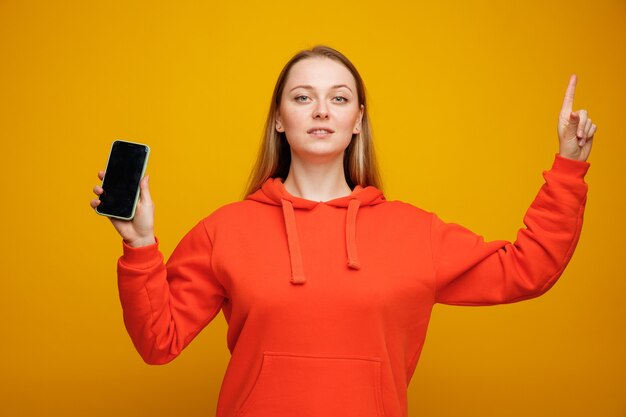Уверенная молодая блондинка женщина, держащая мобильный телефон, указывая вверх