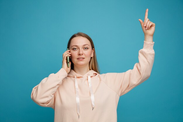 Confident young blonde woman talking on phone pointing up 