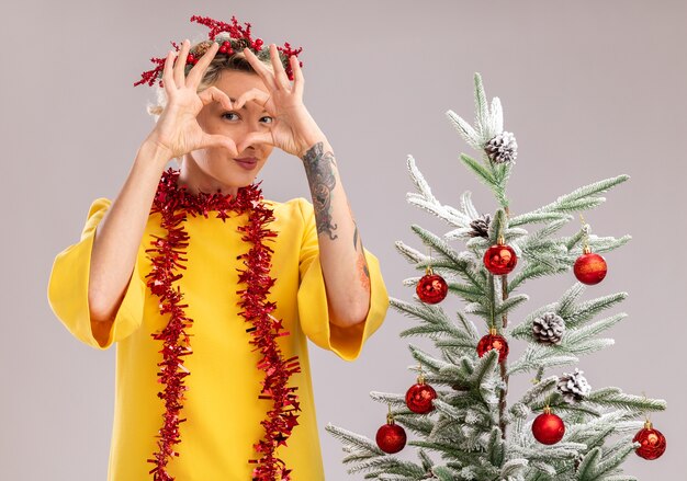 confident young blonde woman wearing christmas head wreath and tinsel garland around neck standing near decorated christmas tree looking  doing heart sign in front of face isolated on white wall