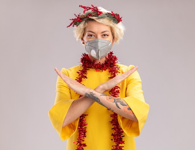 Confident young blonde woman wearing christmas head wreath and tinsel garland around neck with protective mask looking at camera doing no gesture isolated on white background with copy space