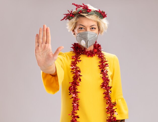 クリスマスの頭の花輪と首の周りに見掛け倒しの花輪を身に着けている自信を持って若いブロンドの女性は、白い背景で隔離の停止ジェスチャーを行うカメラを見て保護マスクと