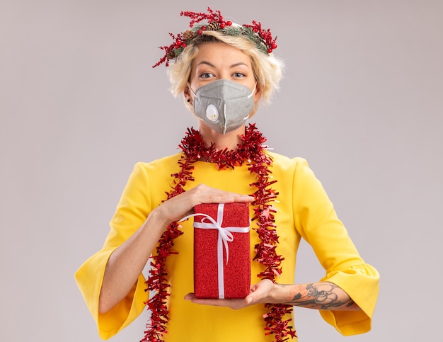 Confident young blonde woman wearing christmas head wreath and tinsel garland around neck with protective mask looking at camera holding gift package isolated on white background