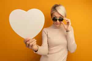 Free photo confident young blonde woman wearing sunglasses grabbing them stretching out heart shape towards front looking at front isolated on orange wall