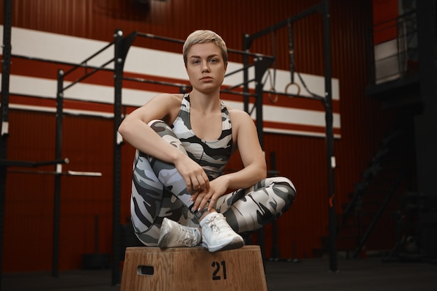 Confident young blonde woman with athletic muscular body and short haircut relaxing in modern gym interior, sitting on wooden platform, looking at camera with serious expression
