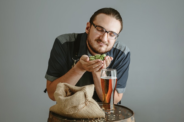 Confident young brewer with self crafted beer in glass on wooden barrel on grey wall