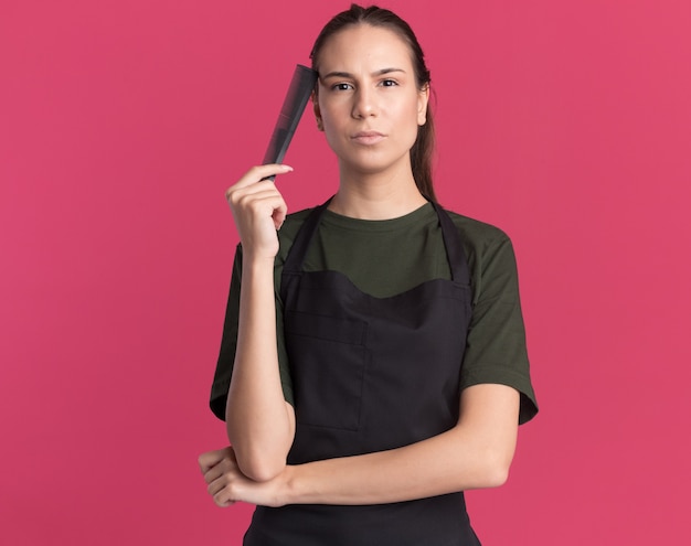 Confident young brunette barber girl in uniform holds comb close to face