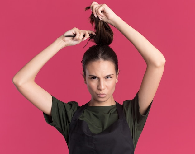 Free photo confident young brunette barber girl in uniform lifts hair and holds comb on pink