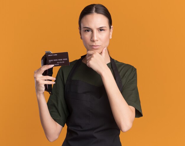 Confident young brunette barber girl in uniform puts hand on chin holding hair clippers and credit card isolated on orange wall with copy space
