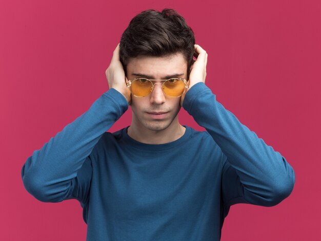 Confident young brunette caucasian boy in sun glasses puts hands on ears looking at camera