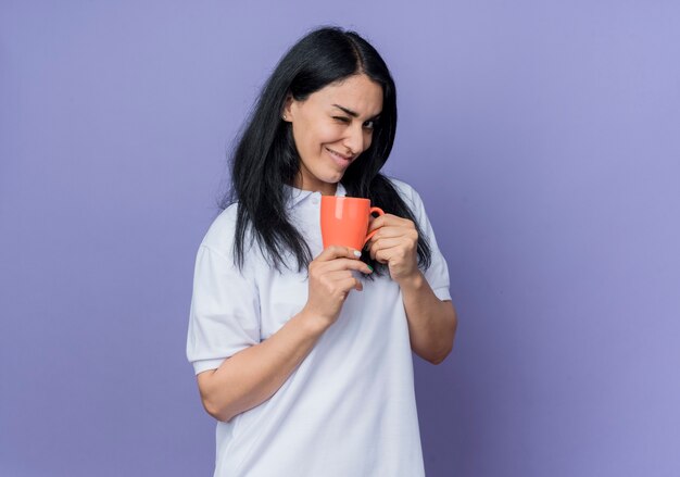 Confident young brunette caucasian girl blinks eye and holds cup looking isolated on purple wall