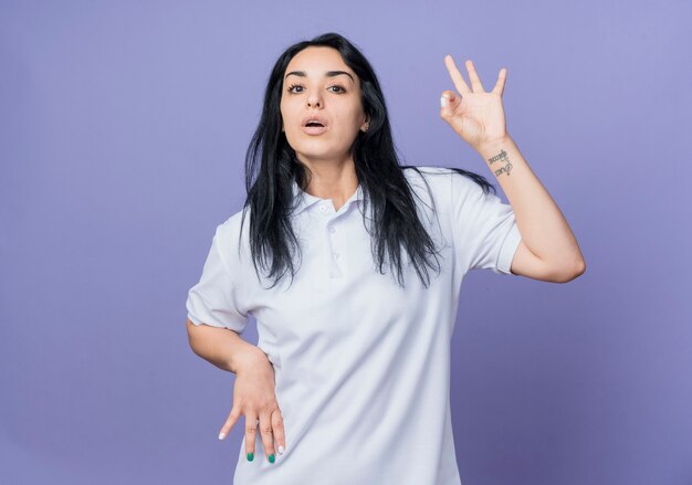 Уверенная молодая брюнетка кавказская девушка жестикулирует знак рукой, изолированные на фиолетовой стене