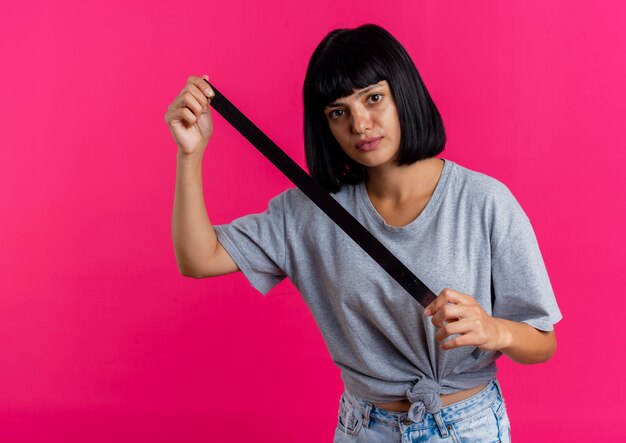 Confident young brunette caucasian girl holds iron ruler looking at camera isolated on pink background with copy space