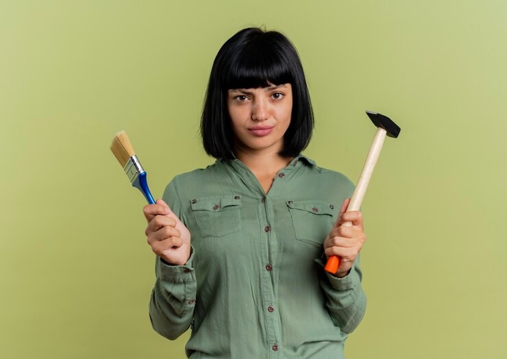 मुफ़्त फोटो confident young brunette caucasian girl holds paint brush and hammer isolated on olive green background with copy space