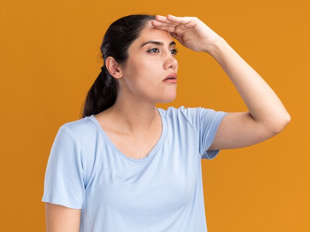 Confident young brunette caucasian girl keeping palm at forehead looking at side on orange