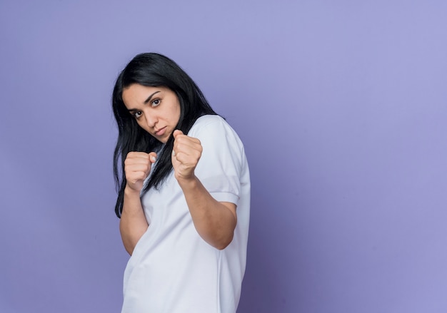 Confident young brunette caucasian girl stands sideways keeping fists ready to punch isolated on purple wall