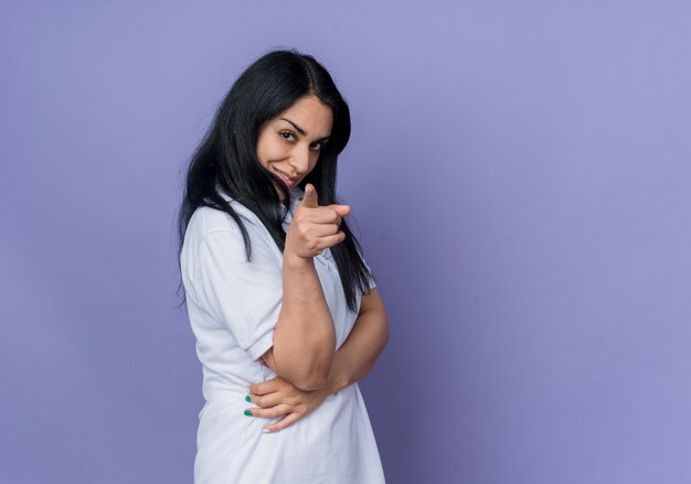 Confident young brunette caucasian girl stands sideways looking and pointing isolated on purple wall