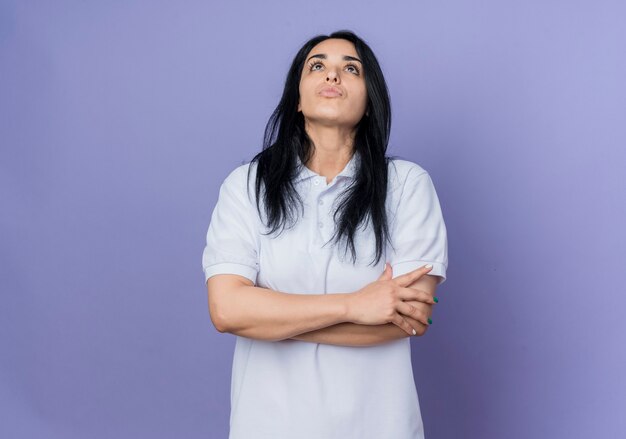 Confident young brunette caucasian girl stands with crossed arms looking up isolated on purple wall