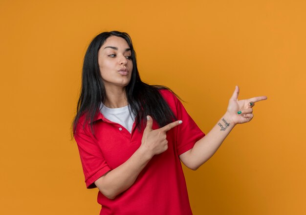 Уверенная молодая брюнетка кавказская девушка в красной рубашке смотрит и указывает в сторону двумя руками, изолированными на оранжевой стене