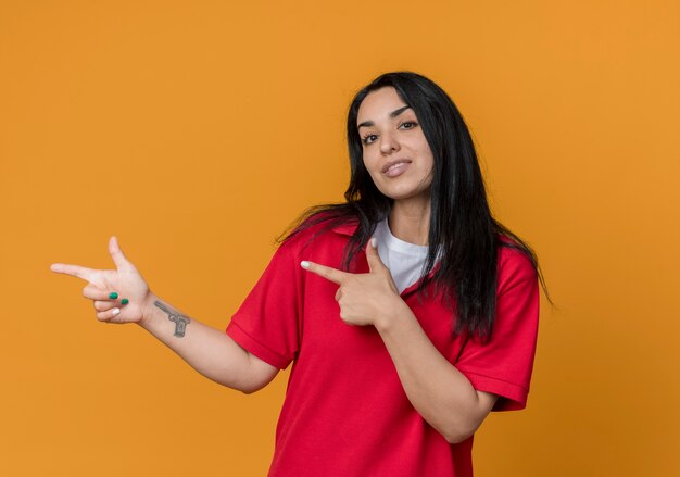 Confident young brunette caucasian girl wearing red shirt points at side with two hands isolated on orange wall