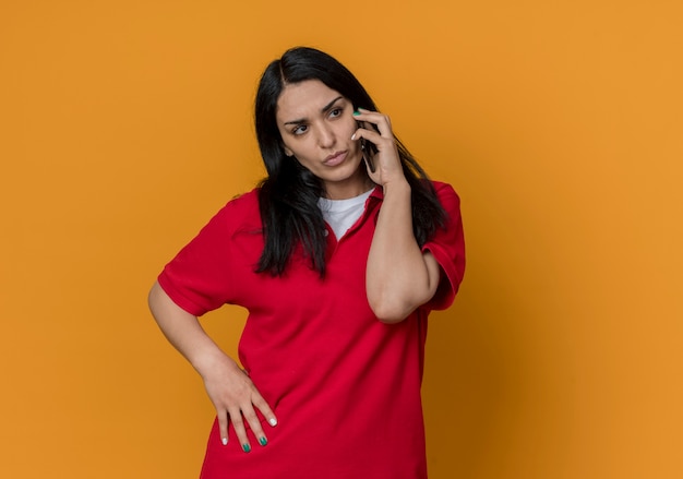 Confident young brunette caucasian girl wearing red shirt talks on phone and looks at side isolated on orange wall