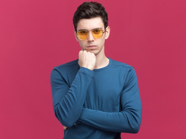 confident young brunette caucasian man in sun glasses puts fist on chin  isolated on pink wall with copy space