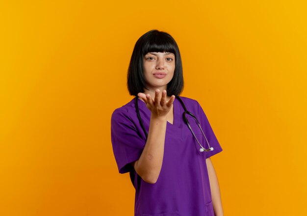 Confident young brunette female doctor in uniform with stethoscope sends kiss with hand 