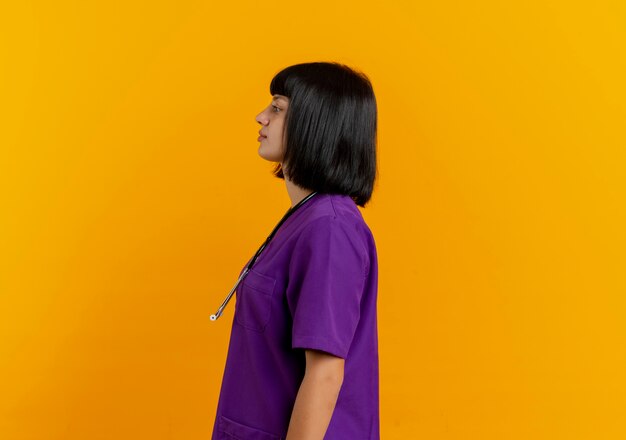 Confident young brunette female doctor in uniform with stethoscope stands sideways