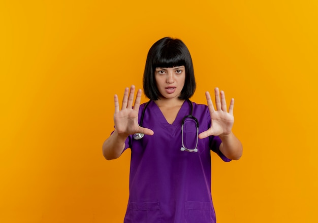 Free photo confident young brunette female doctor in uniform with stethoscope stop hand sign