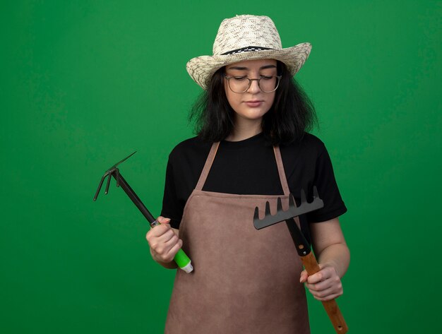 Confident young brunette female gardener in optical glasses and uniform wearing gardening hat holding hoe rake and rake isolated on green wall