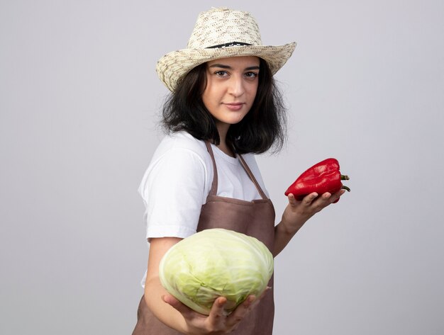 Confident young brunette female gardener in uniform wearing gardening hat holds cabbage and red peppers isolated on white wall