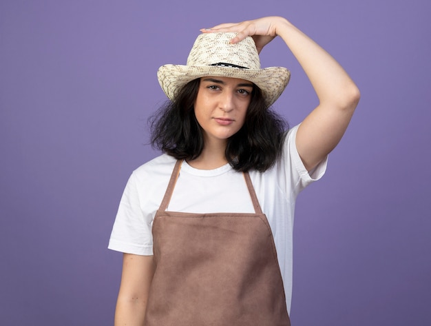 유니폼을 입고 보라색 벽에 고립 된 원예 모자에 손을 넣어 자신감 젊은 갈색 머리 여성 정원사