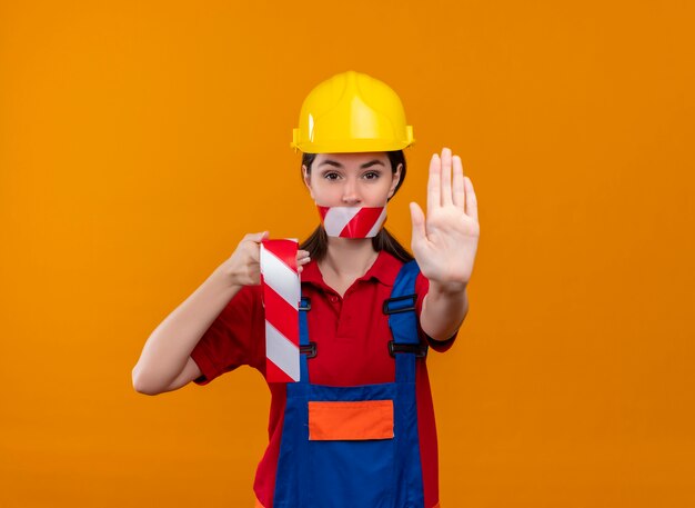 Confident young builder girl mouth sealed with warning tape holds tape and shows stop gesture on isolated orange background with copy space