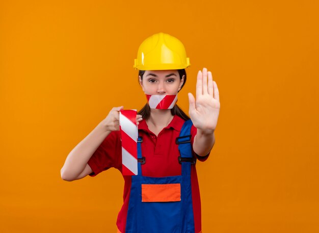 Уверенный в себе рот молодой девушки-строителя, заклеенный предупреждающей лентой, держит ленту и показывает жест остановки на изолированном оранжевом фоне с копией пространства