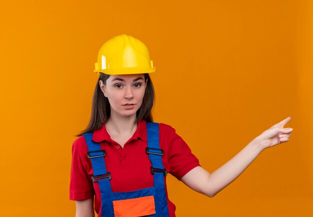 Confident young builder girl points to the side and looks at camera on isolated orange background with copy space