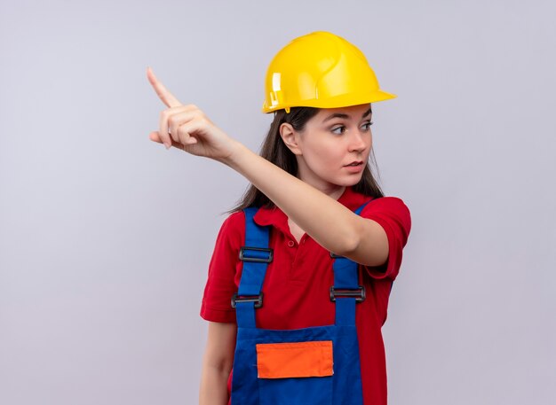 Уверенная молодая девушка-строитель указывает вверх и смотрит в сторону на изолированном белом фоне