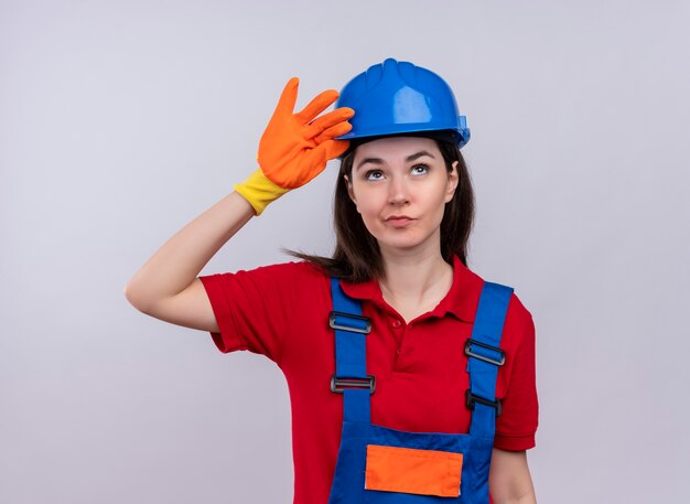 Confident young builder girl puts hand on safety helmet on isolated white background