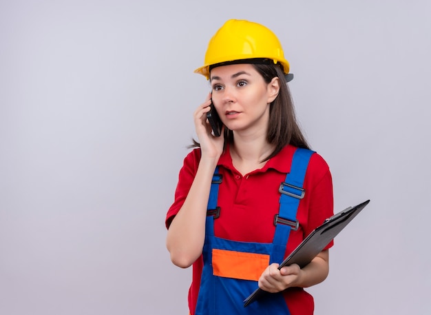 Confident young builder girl talks on phone and holds clipboard on isolated white background with copy space