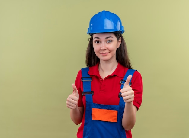 Уверенная молодая девушка-строитель с синим защитным шлемом показывает палец вверх обеими руками на изолированном зеленом фоне с копией пространства