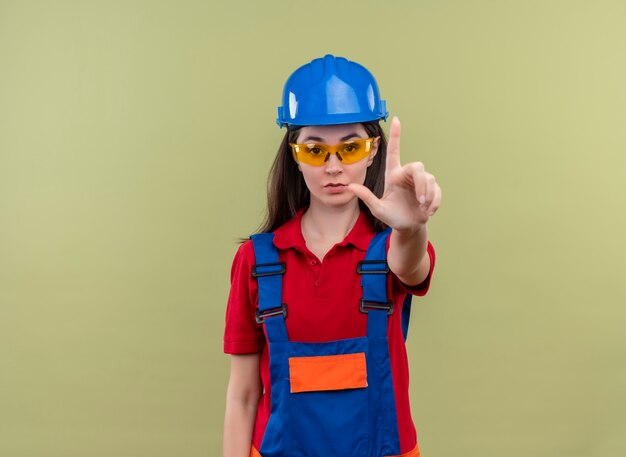 Confident young builder girl with blue safety helmet and with safety glasses holds hand pointing upwards on isolated green background with copy space