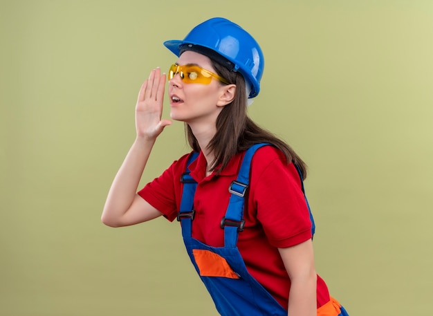 Уверенная молодая девушка-строитель в синем защитном шлеме и в защитных очках смотрит в сторону и делает вид, что зовет кого-то на изолированном зеленом фоне