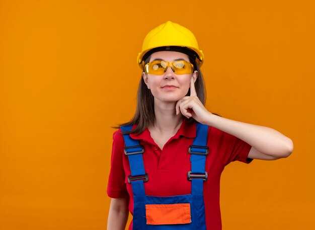 Confident young builder girl with safety glasses puts hand on chin and looks up on isolated orange background with copy space