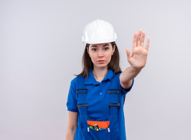 Confident young builder girl with white safety helmet and blue uniform gestures stop on isolated white background with copy space