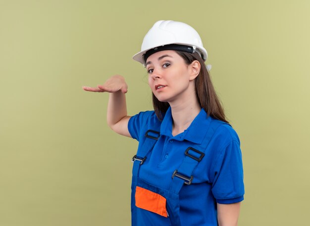 Уверенная молодая девушка-строитель в белом защитном шлеме и синей форме держит руку прямо и смотрит в камеру на изолированном зеленом фоне с копией пространства