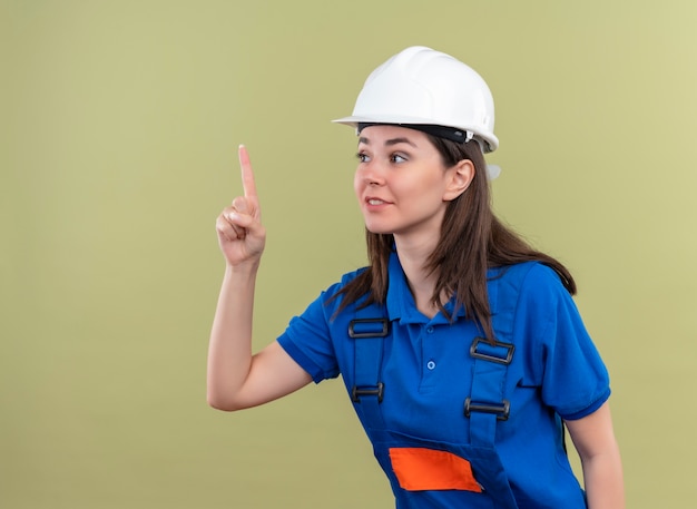 Уверенная молодая девушка-строитель в белом защитном шлеме и синей форме указывает на изолированный зеленый фон с копией пространства