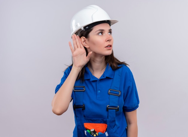 Confident young builder girl with white safety helmet and blue uniform pretends to listen on isolated white background with copy space