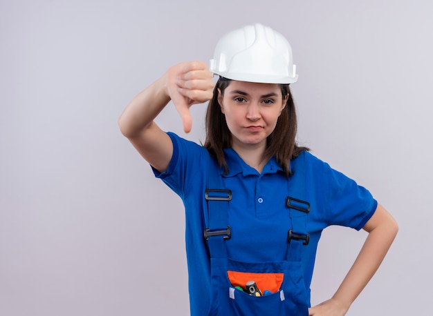Free photo confident young builder girl with white safety helmet and blue uniform thumbs down and puts hand on waist on isolated white background with copy space