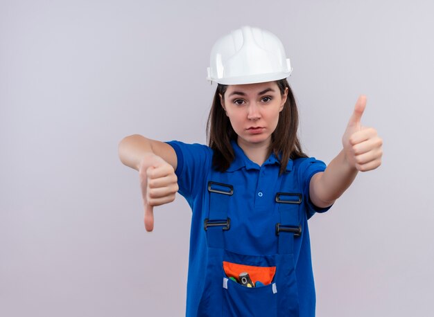 白い安全ヘルメットと青い制服の親指を下に、コピースペースで孤立した白い背景に親指を立てて自信を持って若いビルダーの女の子