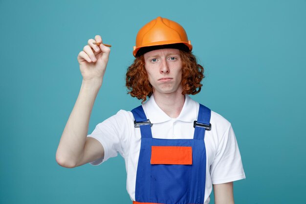 Confident young builder man in uniform holding out pencil to camera isolated on blue background