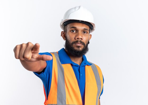 confident young builder man in uniform with safety helmet looking and pointing at front isolated on white wall with copy space