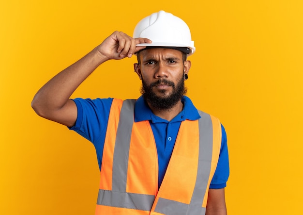 confident young builder man in uniform with safety helmet putting hand on helmet isolated on orange wall with copy space